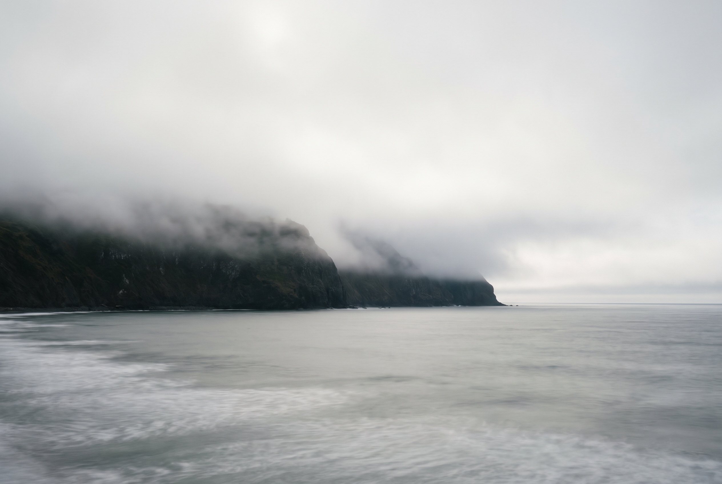 Fog rolling over a rugged coastal cliff at dawn, the sea smoothed to a blur by a long exposure