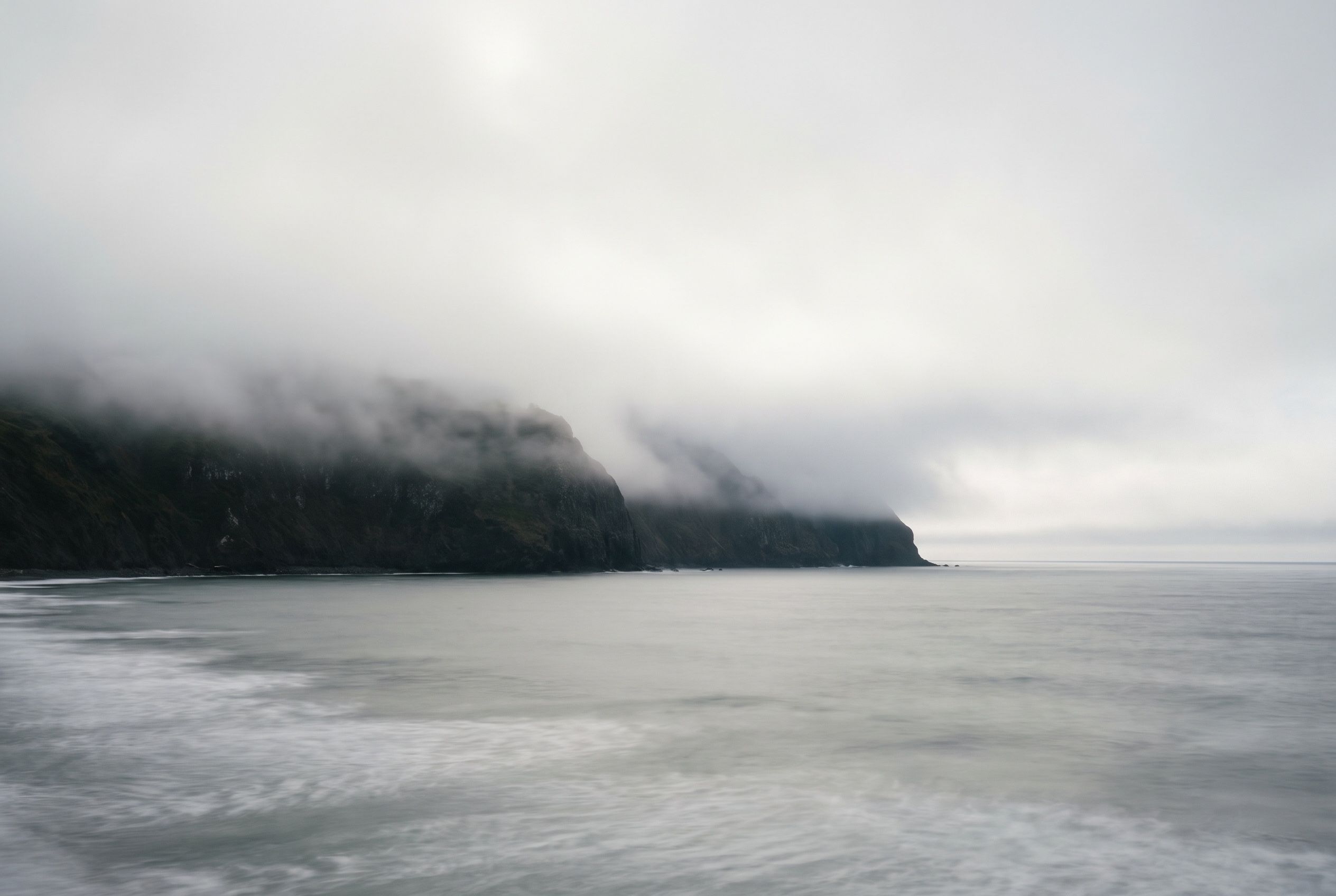 Fog rolling over a rugged coastal cliff at dawn, the sea smoothed to a blur by a long exposure